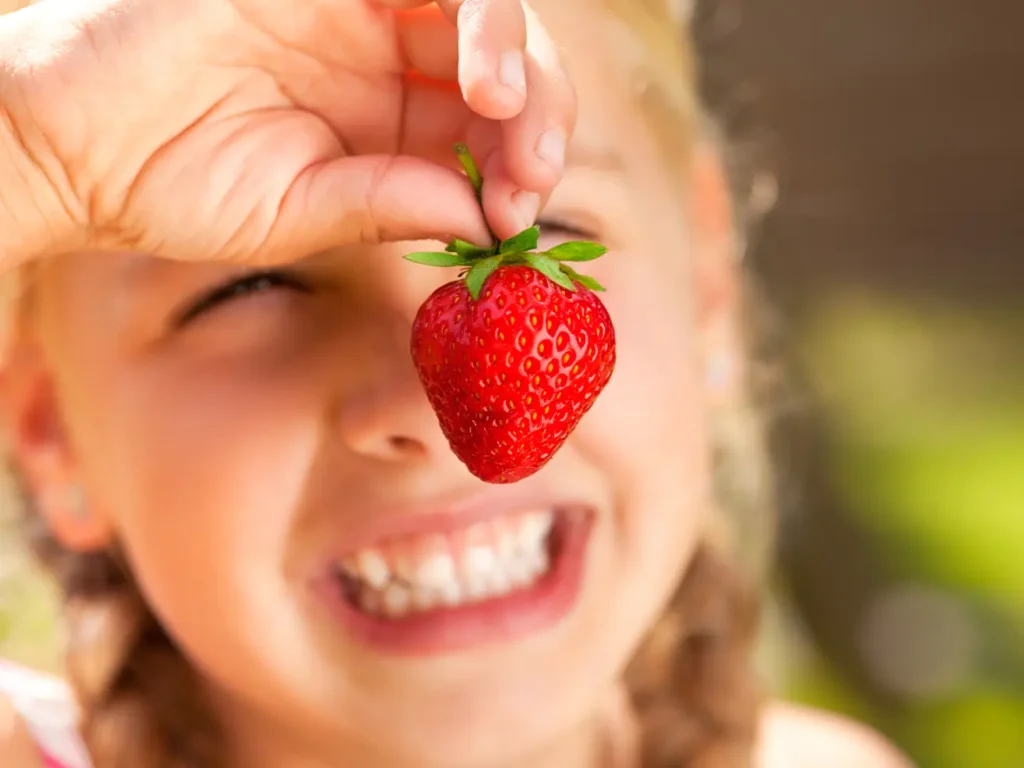 Kleines lachendes Mädchen mit einer Erdbeere vor dem Gesicht